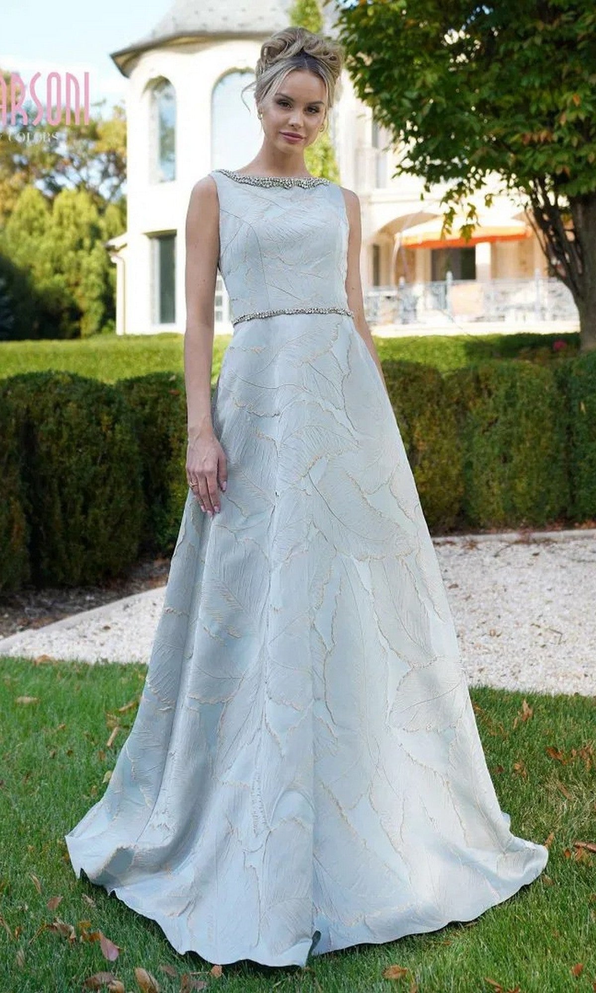 Outdoors on grass, a woman wears the Marsoni MV1224 Long Formal Dress—a sleeveless, light blue designer gown with jacquard print and beaded trim—with a house and greenery in the background.
